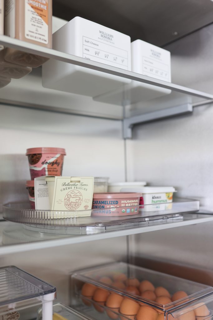 Refrigerator shelf with a labeled cheese bin, a turntable of dips and hummus, and an egg bin on the shelf below.